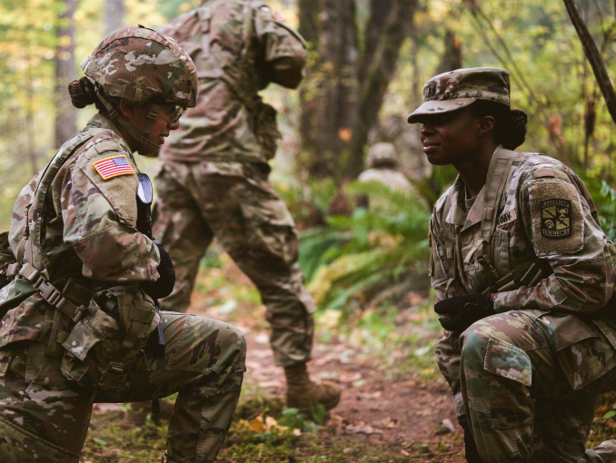 Two female ROTC cadets, one holding a rifle, kneeling in a forest