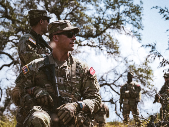 A Lieutenant wearing combat uniform and sunglasses kneeling and holding a rifle in a field on a sunny day