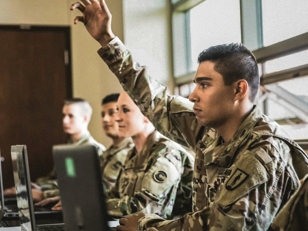 A row of soldiers in combat uniform sit in front of laptops in a classroom as one soldier raises his hand