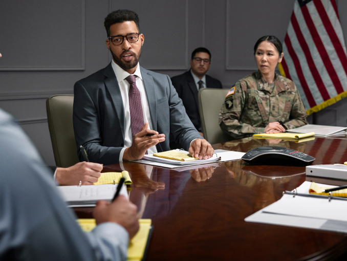 A man in a navy suit sitting next to a female Soldier in combat uniform in a board room