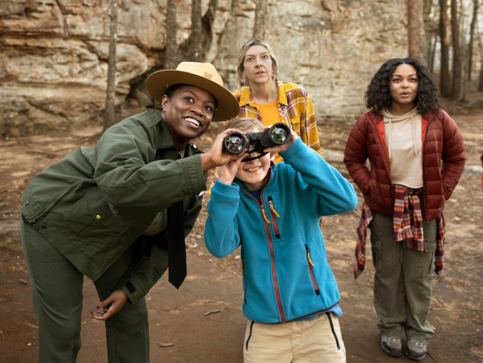 A female park ranger assisting a young boy with binoculars in a forest preserve