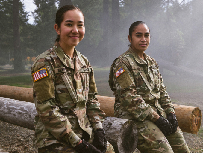 Two female ROTC cadets sitting outside against a wooden obstacle