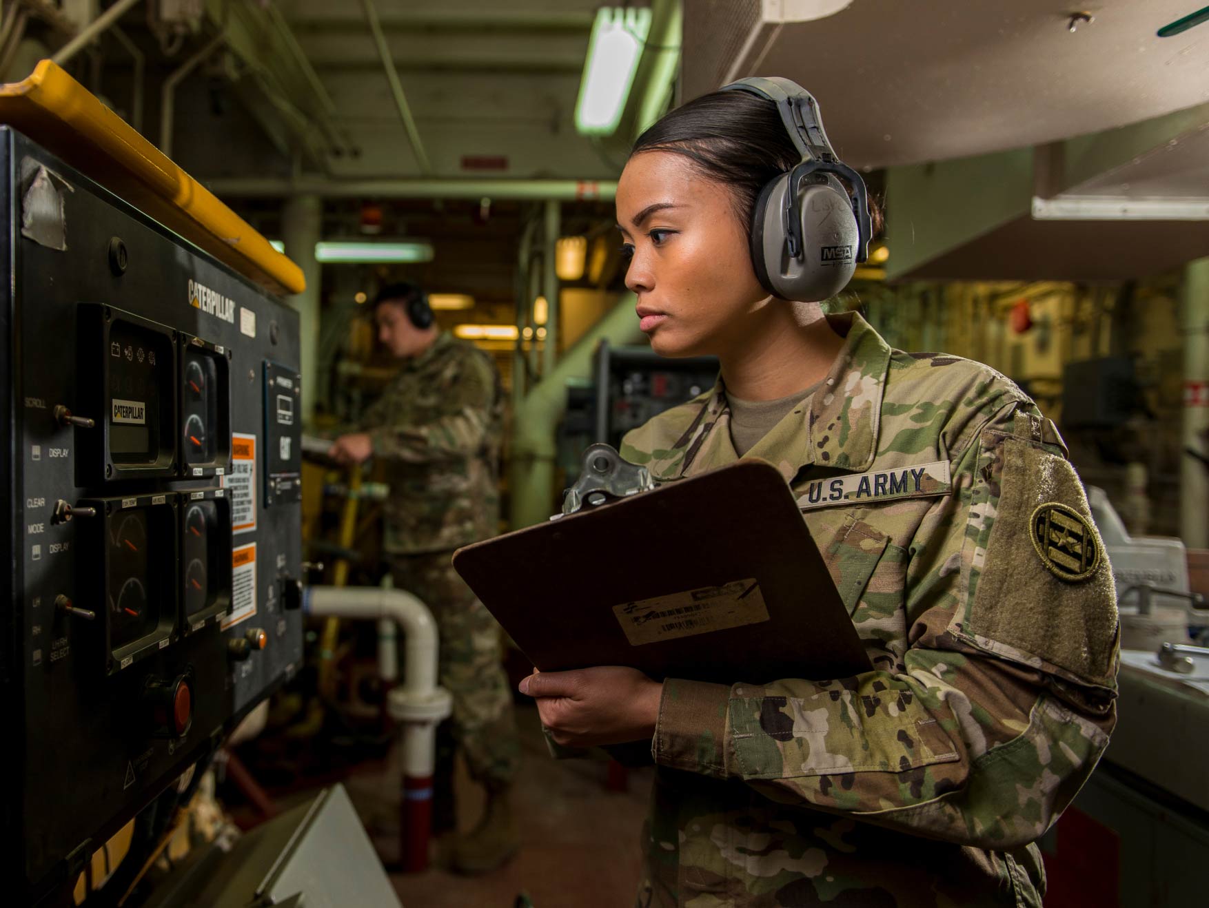 A female Soldier wearing headphones reading gauges on an Army watercraft