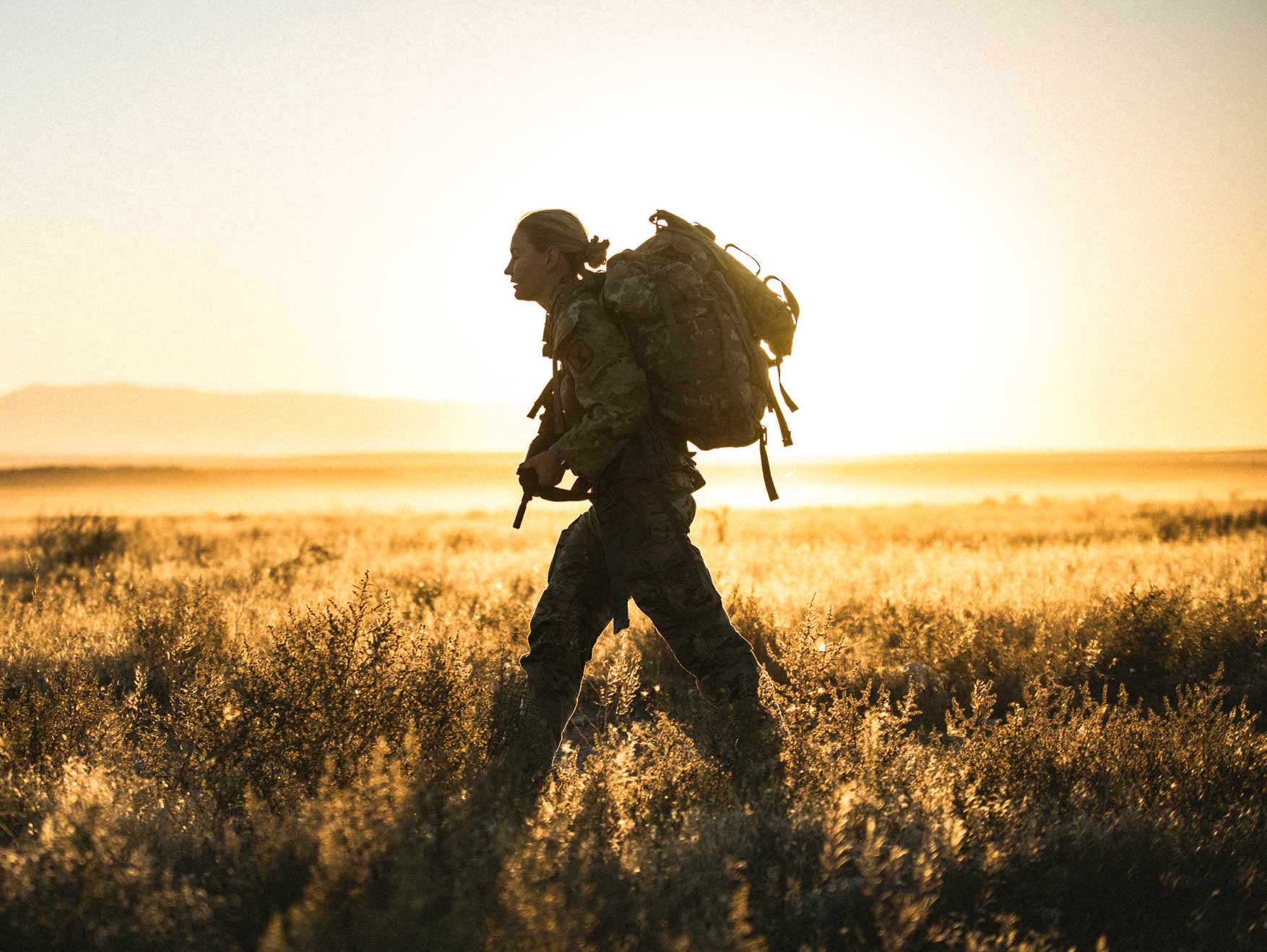 A female Soldier wearing a rucksack walking through a field carrying a rifle