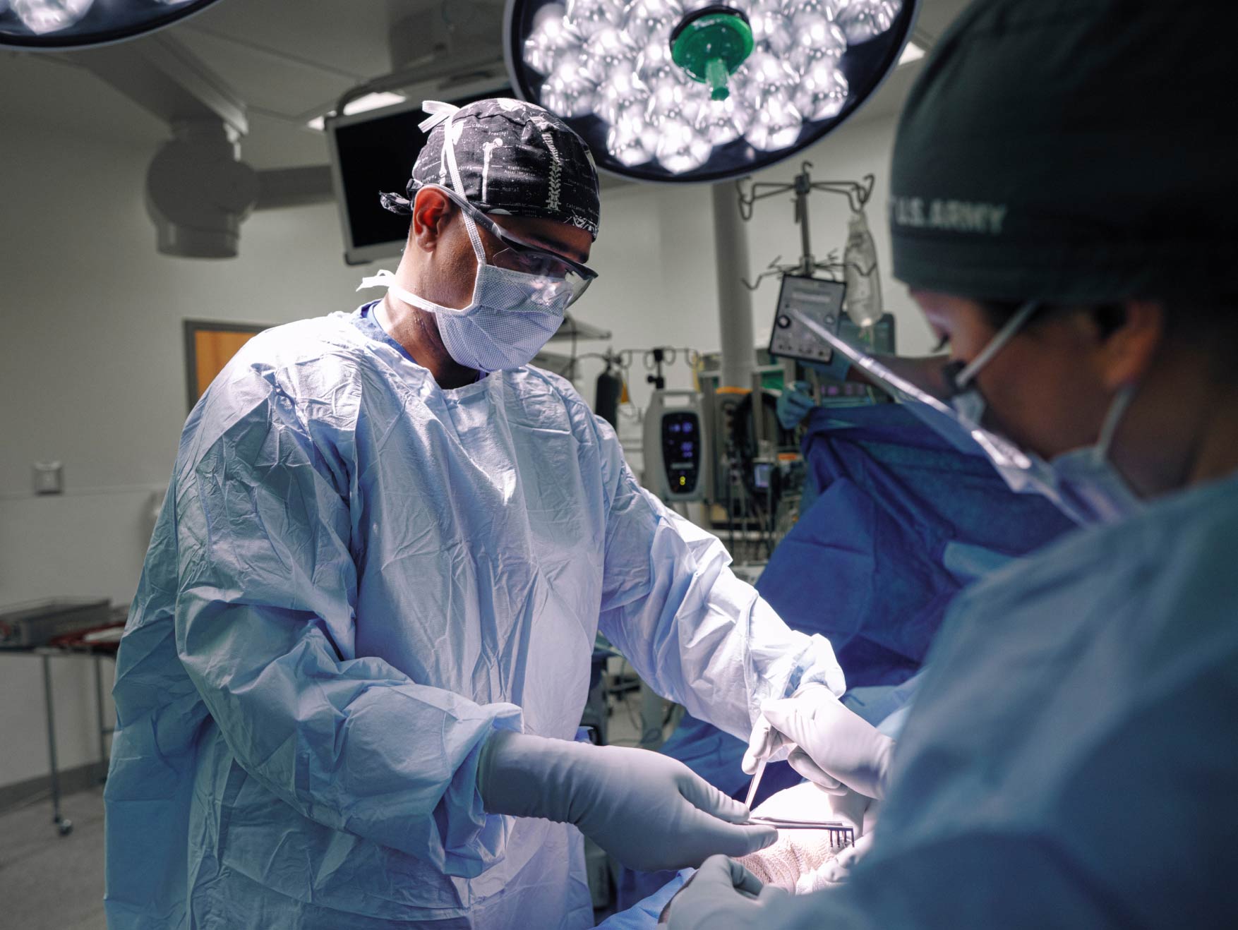 An Army Surgeon wearing a gown, mask, and army cap with patient